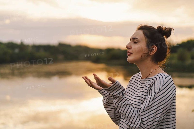 A serene moment captured as a woman praying in a striped sweater enjoys a peaceful sunset by the lake. Her expression reflects calmness and hope in the golden light