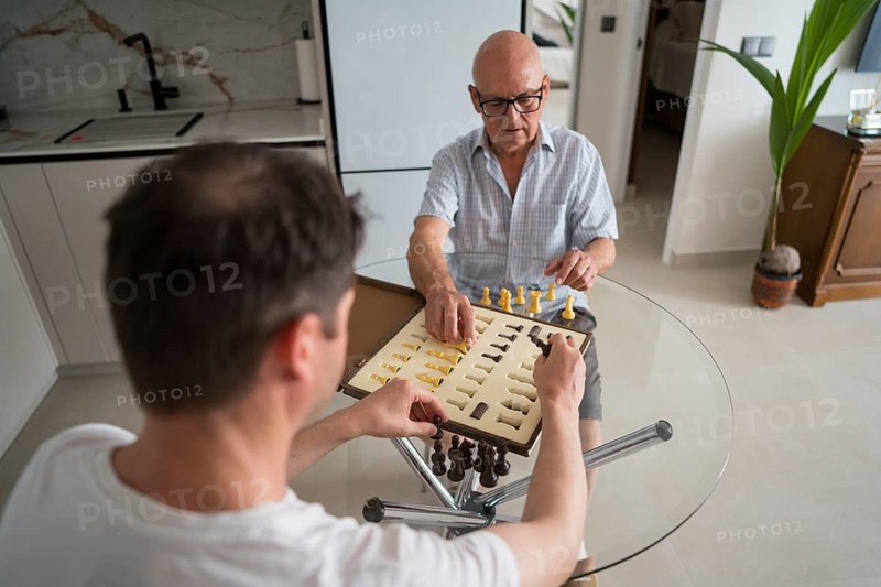 Back of unrecognizable adult son and elderly father enjoying a chess game at home, showcasing quality time and bonding across generations in a cozy setting
