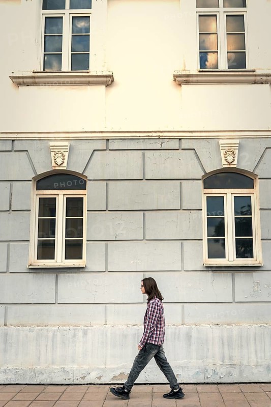 A teenage boy with long hair walks along a historic building with classic architectural details, wearing a plaid shirt and jeans on a paved sidewalk