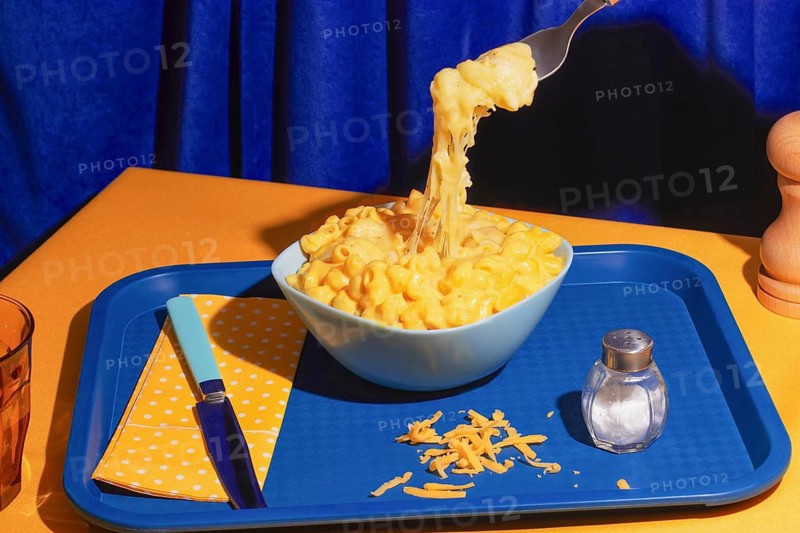A close up of creamy mac and cheese in a blue bowl on a colorful table setting. A fork lifts the cheesy pasta, with a salt shaker and napkin nearby, creating a vibrant scene