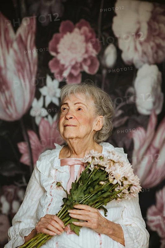 An elderly woman in a studio holds a bouquet of flowers, looking serene. She stands against a background of large floral patterns, creating a timeless portrait