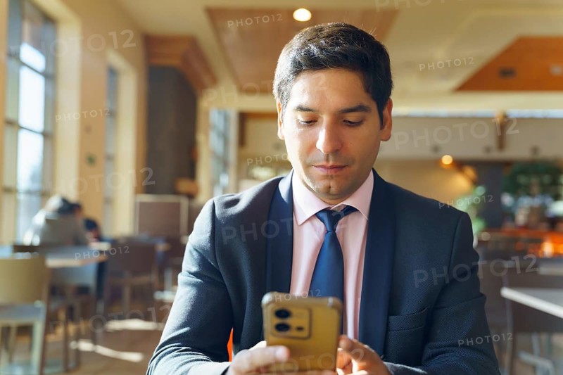 Young Chilean businessman in a contemporary cafe, wearing a suit and using a smartphone. Sunlight filters through large windows, creating a professional yet relaxed atmosphere