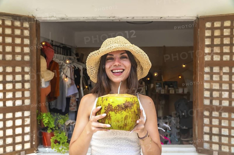 Young woman with straw hat drinking fresh coconut water in front of a shop in siargao island, philippines, during summer holidays