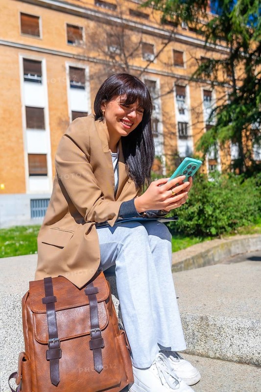 Female university student sitting on campus steps, smiling and using her smartphone with a backpack beside her on a bright, sunny day outdoors, enjoying studying and social connection