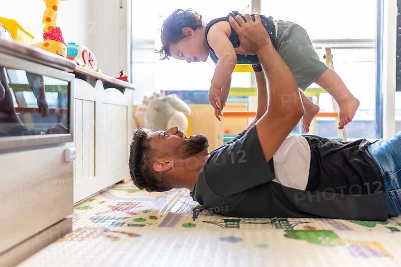 Father lying on a playmat, joyfully lifting his toddler son into the air, creating a strong bond and sharing a happy moment together in a bright home environment