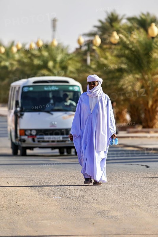 A man in traditional dress walks on a road through a desert landscape with palm trees and a bus, Traditional Tuareg walking across the road while a bus is coming, Sahara Algeria