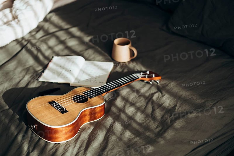 A ukulele rests beside an open notebook and a coffee mug on a softly sunlit bed This cozy scene evokes a sense of relaxation, creativity, and leisurely comfort