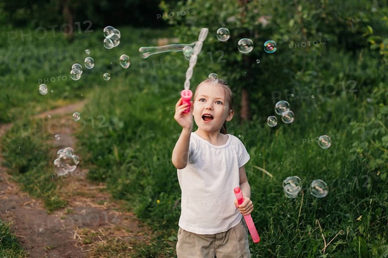 A joyful little girl plays with soap bubbles in a lush green outdoor setting. Her expression of happiness captures the essence of a carefree summer day in childhood
