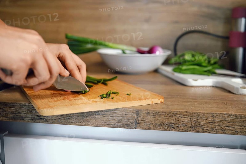 Close-up view of trimming ends of green beans with knife on chopping board. Fresh vegetables are on counter. Bowl with other vegetables in background. Kitchen setting shows preparation for meal