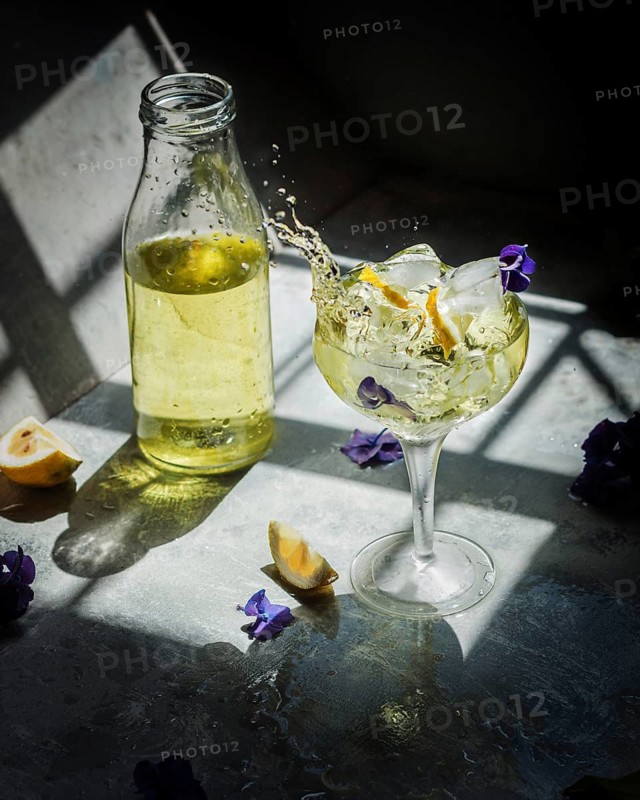 Lemon liqueur in a glass, with ice cubes and lemon slices, splashes dramatically in sunlight. Nearby, a bottle and flowers complete the vibrant setting