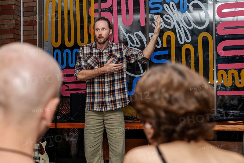 A man with a beard and plaid shirt speaks to an audience in a colorful venue. Two people with short hair listen attentively from the foreground