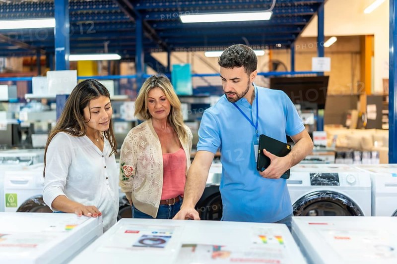Salesman helping female customers compare washing machines and appliances, discussing features and energy labels in a home improvement store showcasing customer service and shopping experience