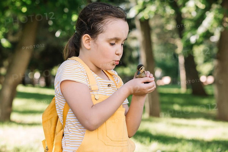 In a sunny, green park, a young girl in a yellow outfit holds a fluffy duckling, showcasing a moment of innocence and tenderness as she smiles at the cute bird