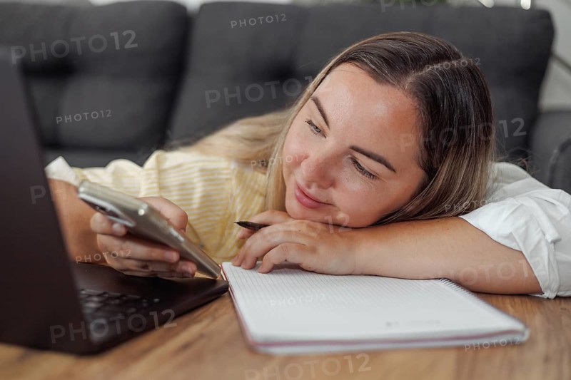 Young Caucasian woman with brown hair lying on a couch using a smartphone with a laptop and notebook on the table