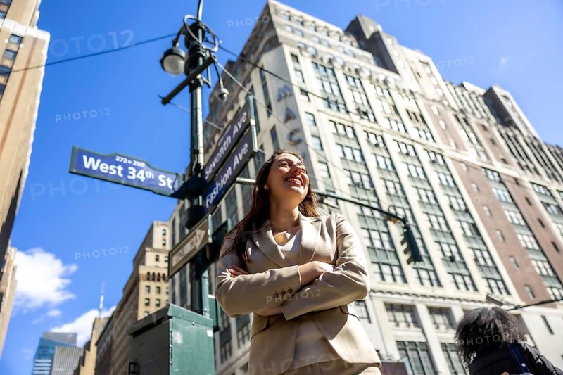 Confident businesswoman standing with arms crossed, smiling brightly while looking up against a backdrop of towering buildings and street signs in the vibrant heart of new york