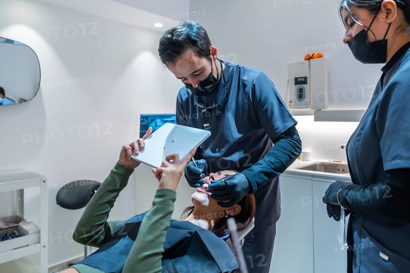 Dentist showing teeth whitening results to a patient on a digital tablet, assisted by a dental assistant, in a modern dental clinic