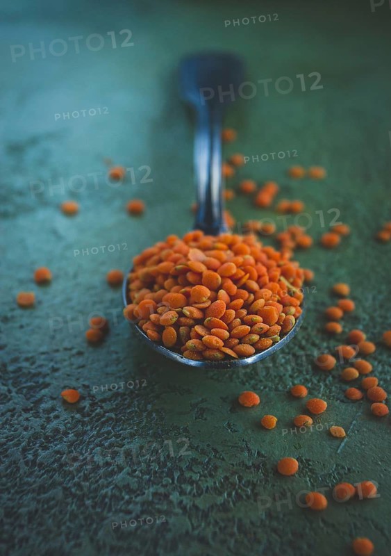 A spoon rests on a textured surface, holding a mound of vibrant orange lentils. Some lentils scatter around it, showcasing their lively color and natural shape during food preparation.