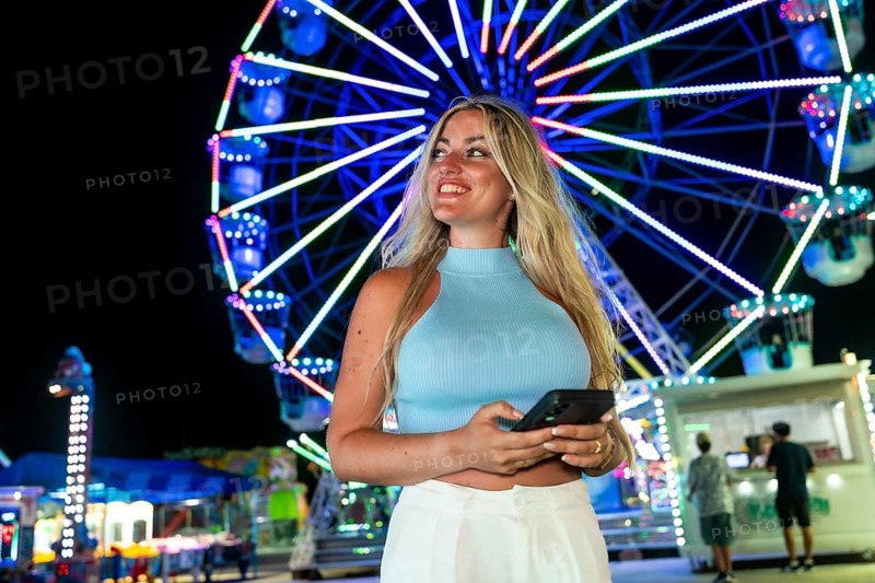Young woman with blonde hair smiling and looking away while holding a smartphone with a brightly lit ferris wheel spinning in the background at a lively amusement park during the evening