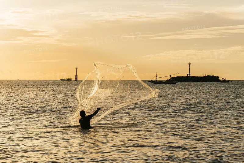 A silhouette of a fisherman casting a net into the ocean during a golden sunset. Boats are visible in the distance, creating a serene and traditional maritime scene