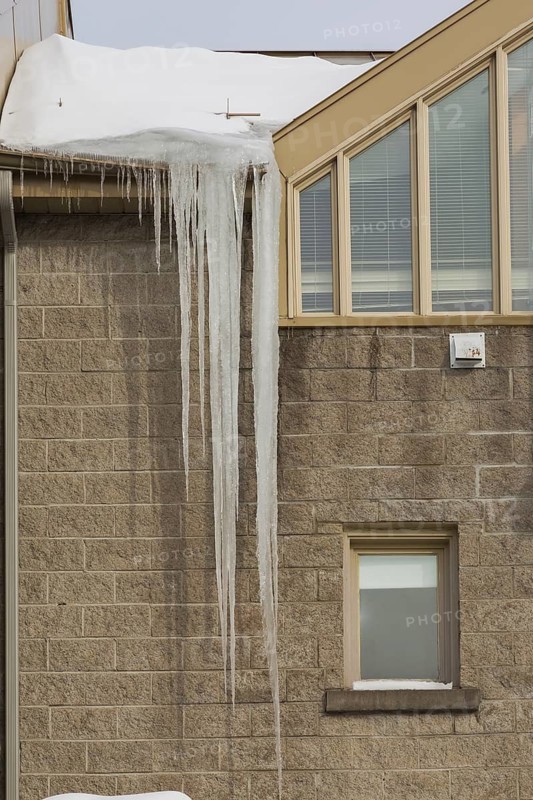 Long icicles hanging from roof edge of light brown cut stone two storied building with dameged gutter in winter, Quebec, Canada