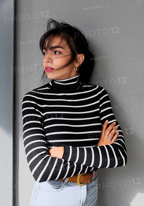 A woman in a black and white striped turtleneck sweater leans against a gray wall. She crosses her arms and gazes to the side, conveying a thoughtful mood