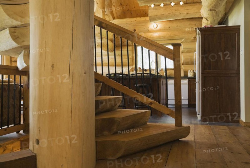 Twisting staircase with wooden and black wrought iron handrail and brown stained half log steps attached to large supporting central tree trunk post leading to mezzanine from hallway inside handcrafted Eastern white pine Scandinavian log cabin home, Quebec, Canada