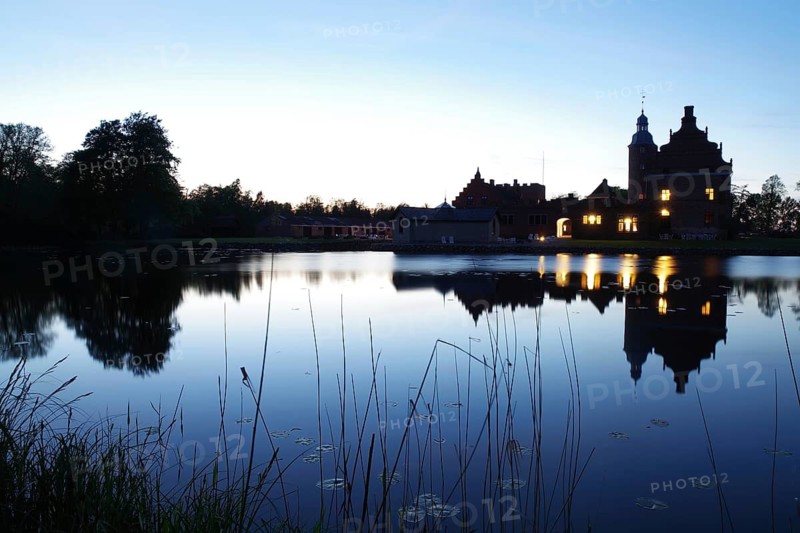 A castle at dusk reflected in a quiet lake with glowing windows, Broholm, Funen, manor house route, Denmark