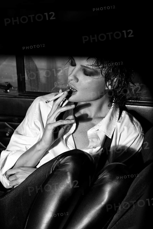 Elegant woman sitting in a car, holding a cigarette. Dramatic lighting highlights her stylish attire, including a white shirt and leather pants, creating a cinematic effect