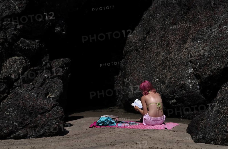 Girl, woman, from behind, in bikini, hair mauve, purple, sitting between rocks on bath towel, pink, with aura cards, reading book about magic wands, fortune teller, sand, sandy beach, beach, Kynance Cove, Mount's Bay, Lizard Peninsula, peninsula, Cornwall, England, Great Britain, Great Britain