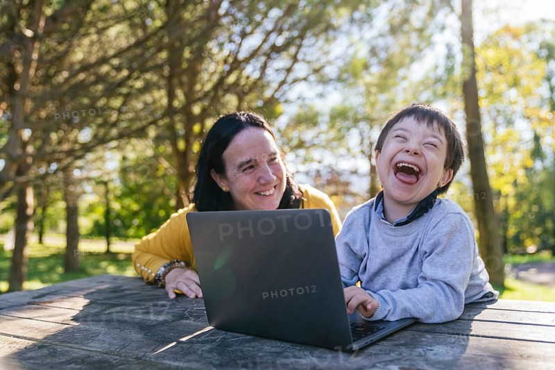 A joyful moment captures a boy with Down syndrome and his mother engaging with a laptop outdoors. The child is laughing heartily, showcasing the blend of technology and nature in education and play