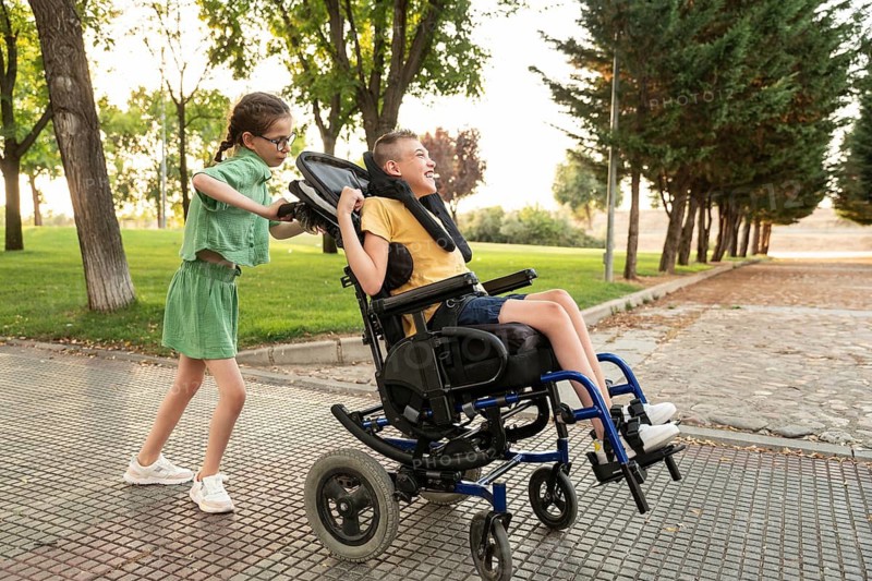 Two friends, a boy with cerebral palsy in a wheelchair and a girl with a tracheostomy, share a joyful moment outdoors on a sunny day, highlighting friendship and inclusion