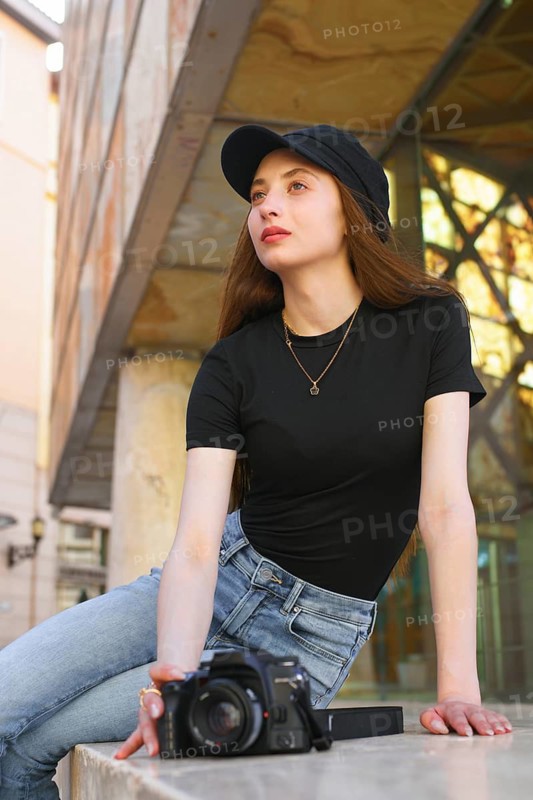 A woman in casual attire, with long hair and a cap, leans against a city building holding a camera. The urban backdrop reflects a modern professional lifestyle
