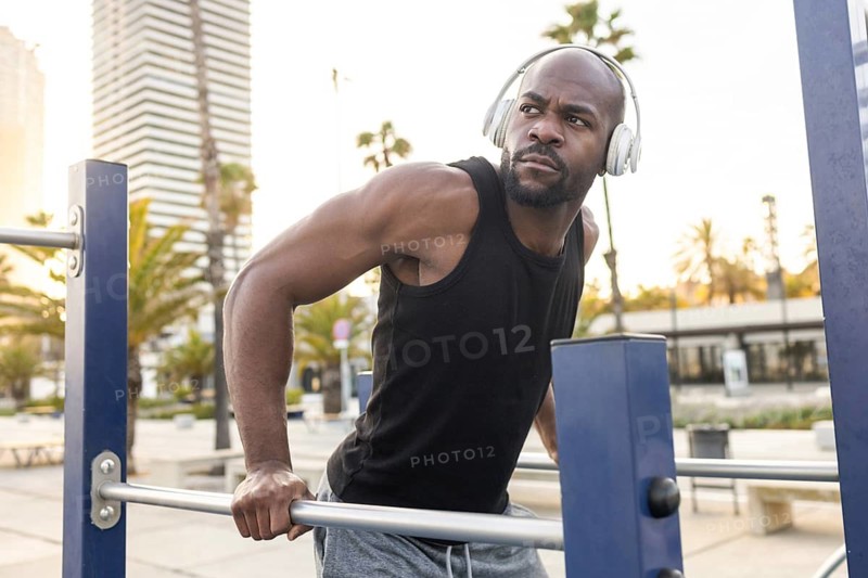 Cuban man in a black tank top works out at an outdoor fitness park, gripping parallel bars. He wears headphones, focused on his exercise routine. Cityscape in the background