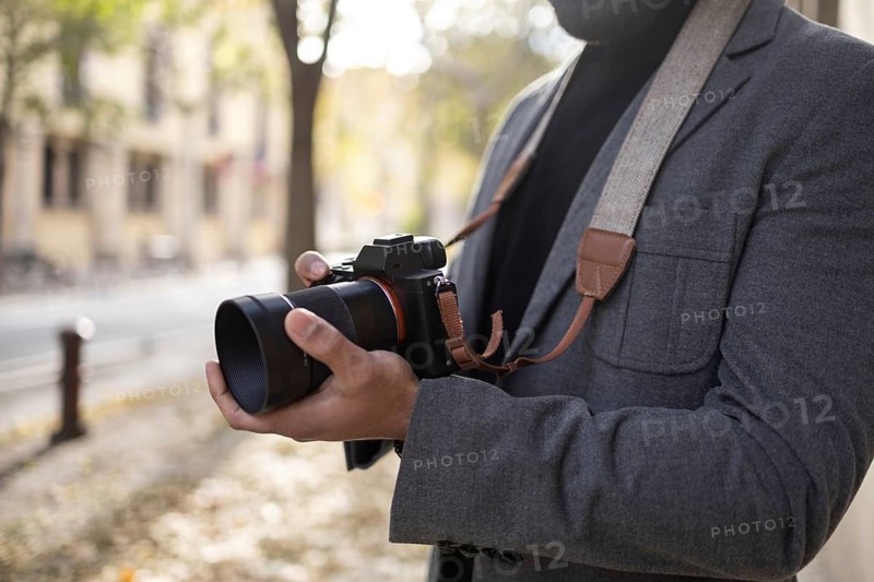 Cropped unrecognizable man, dressed as a photographer with a stylish gray jacket and a camera strap, holds a digital camera, focusing intently on capturing the perfect autumnal street scene