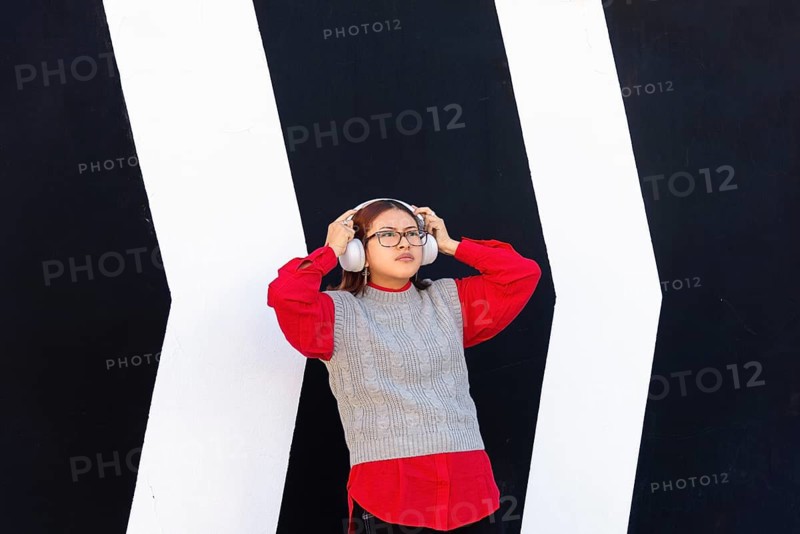 A Latina woman is standing in front of a striking black and white geometric wall, wearing a red shirt and a grey vest She is listening to music through large headphones