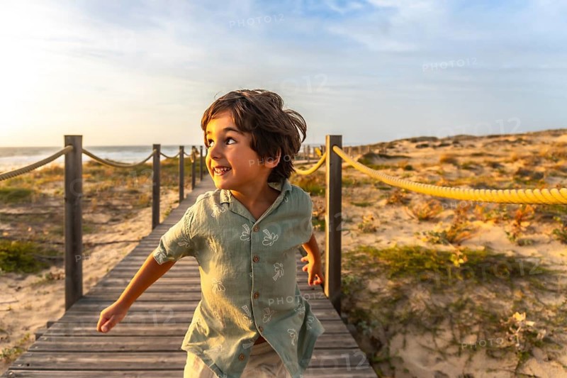Happy young boy running along a wooden boardwalk over sand dunes at sunset in seignosse, france, enjoying carefree summer beach vacation and golden hour freedom on the coast