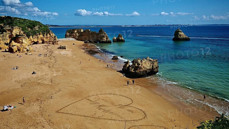 A big heart is drawn in the sand on the beach, International Women's Day, with rocks in the background and people in the sand, Praia Dona Ana, Lagos, Algarve, Portugal
