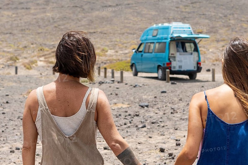 Two lesbians women walk toward a turquoise camper van on a desolate, rocky landscape, embodying exploration and adventure. Their backs are to the camera, with the van in the distance