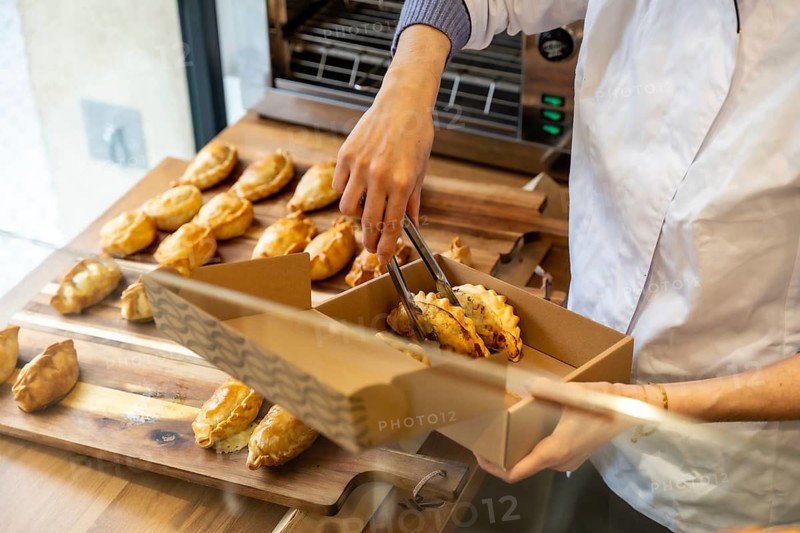 Baker's hands using tongs to carefully place traditional empanadas from a wooden serving board into a recyclable cardboard take out box, preparing a fresh order for a customer in a bakery or shop