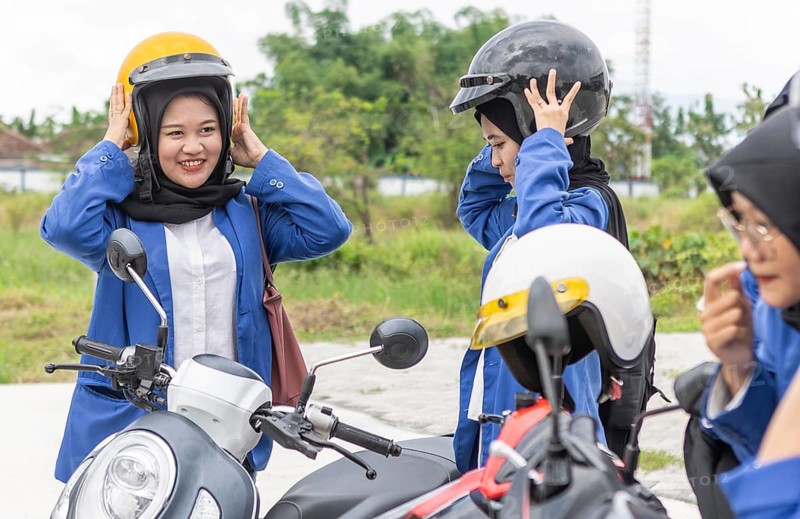 A group of Asian Muslim students prepare for their journey back to school, wearing helmets. hijabs and backpacks, as they chat and get ready to ride motorcycles on a school day