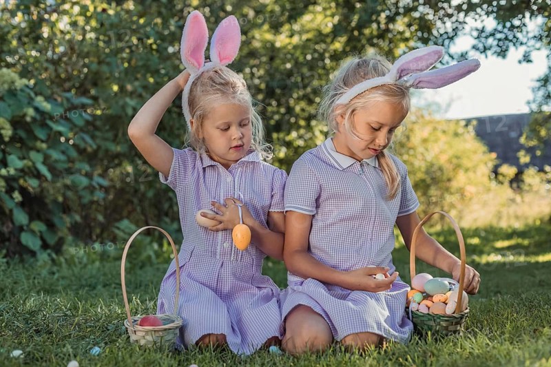 Two young girls, wearing bunny ears and dresses, search for colorful Easter eggs in a sunny garden setting. Each holds a basket filled with eggs, enjoying the festive activity