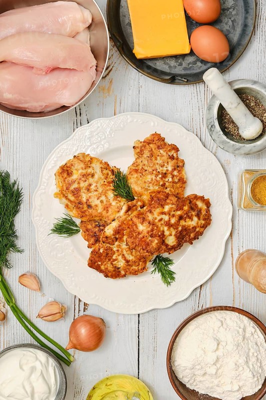 Chicken fritters are being prepared in the kitchen. Nearby are bowls containing chicken breasts, eggs, cheese, flour and spices. Fresh herbs and vegetables are also present