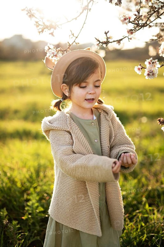 A young girl, wearing a beige hat and sweater, admires almond blossoms on a sunny spring day