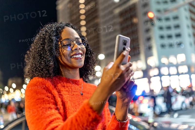 Smiling woman browsing social media on mobile phone in urban environment with bokeh lights
