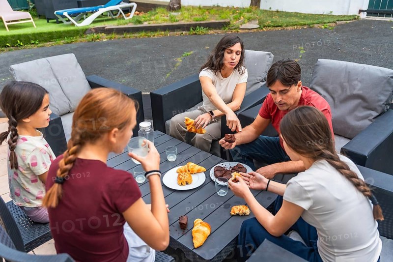 Family gathered around a patio table enjoying pastries and drinks, sharing relaxed conversation and laughter for a casual summer backyard moment of togetherness and leisure