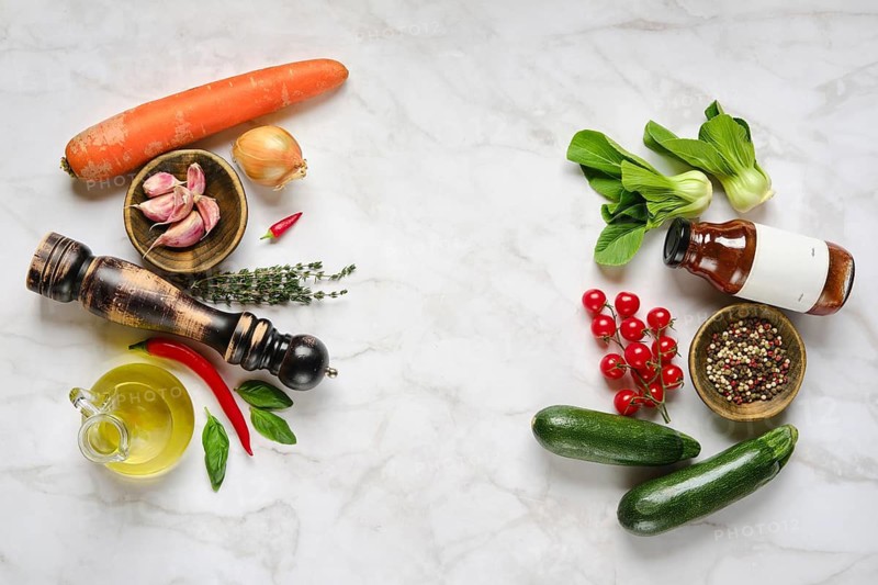 A variety of fresh vegetables, herbs, and condiments are beautifully arranged on a marble countertop, highlighting the colors and textures. Ingredients include zucchini, tomatoes, garlic, and onion.