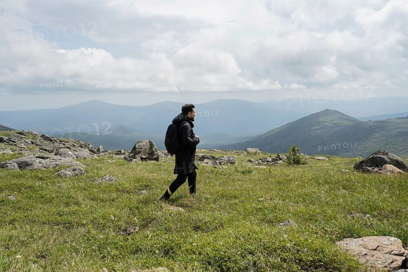 A lone hiker walks across a grassy mountain terrain, surrounded by rocky outcrops and distant peaks under a cloudy sky, embodying adventure and natural beauty