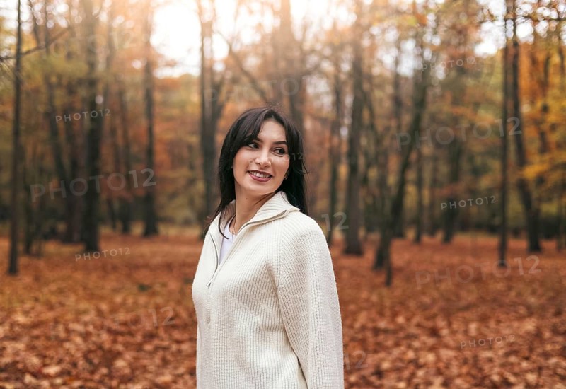 A woman wearing a cozy sweater stands in a forest filled with vibrant autumn foliage. The ground is covered in orange leaves, creating a warm, serene atmosphere