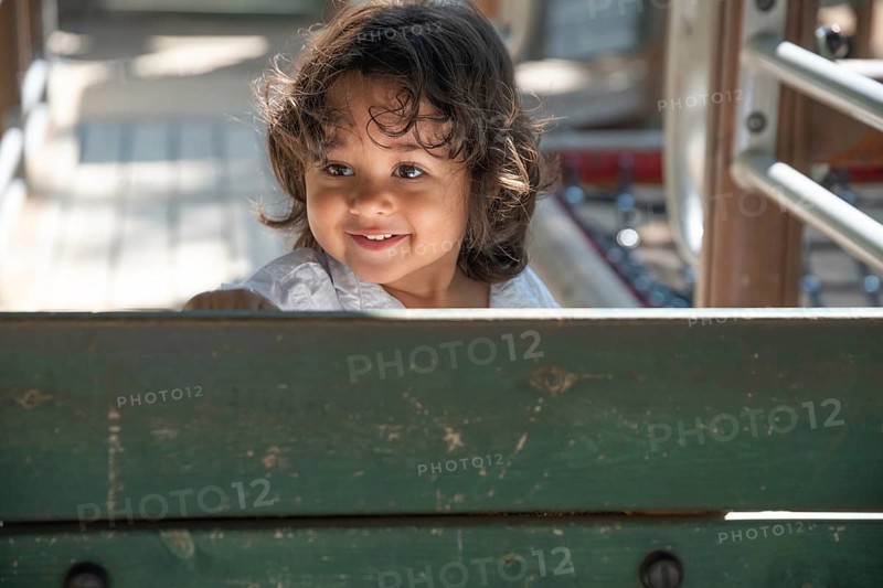 A young boy joyfully peers over a wooden fence, enjoying a sunny day at the playground. The scene captures a moment of pure childhood happiness and outdoor fun