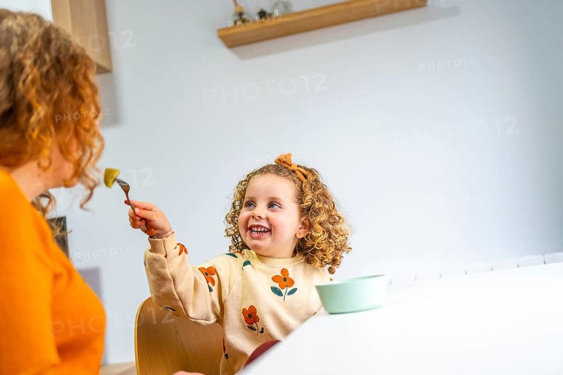 Playful and smiling caucasian school girl eating at home next to her mother